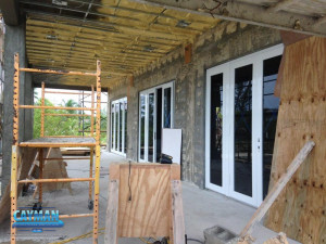 Scaffolding and wooden boards are seen on the outside deck while spray foam insulation is installed on the ceiling.
